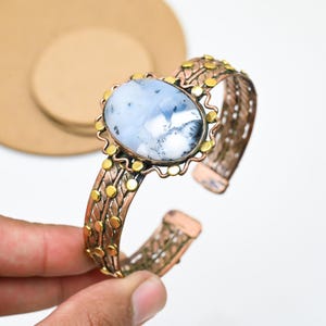 Könnte beinhalten: Ein kupferfarbenes Armreif-Armband mit einem ovalen, hellblauen und weißen Stein. Der Stein ist in einer goldfarbenen Lünette gefasst. Das Armband hat ein dekoratives Muster aus goldfarbenen Punkten und gewebtem kupferfarbenem Metall.