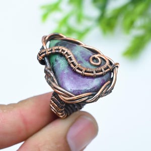 Könnte beinhalten: Ein Ring mit einem tropfenförmigen Stein in gesprenkeltem Violett und Grün, eingefasst in eine dekorative Kupferdrahtwicklung. Der Metalldraht ist um den Stein gewickelt und bildet die Ringschiene, mit einem dunklen, oxidierten Finish.