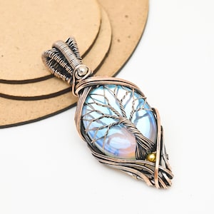 May include: A wire-wrapped pendant featuring an oval, iridescent light blue cabochon stone. The copper wire is intricately coiled to form the shape of a tree over the stone, with a small gold-toned bead at the base and a coiled bail at the top.