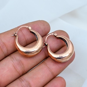 May include: A pair of small, thick hoop earrings with a polished copper finish. The earrings feature a rounded, chunky design with a hinged closure mechanism at the top, showcasing a warm, metallic reddish-brown tone throughout the surface.