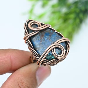 Könnte beinhalten: Ein tropfenförmiger Ring mit einem blauen und braunen Stein, der mit kunstvollem Kupferdraht umwickelt ist. Der Metalldraht ist zu dekorativen Spiralen und engen Bändern geformt, die den Edelstein fixieren und einen detaillierten Rahmen bilden.