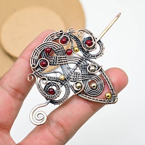 Könnte beinhalten: Eine handgefertigte Haarnadel aus Kupfer mit aufwendiger Drahtarbeit. Die Nadel hat ein wirbelndes Design mit eingebetteten roten und goldfarbenen Perlen. Die Nadel wird in einer Hand gehalten, was die detaillierte Handwerkskunst und den einzigartigen Stil zeigt.