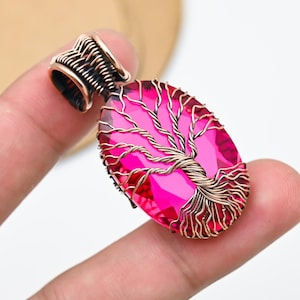 Könnte beinhalten: Handgefertigter Anhänger mit einem leuchtend rosa Edelstein in ovaler Form. Der Edelstein ist in einem kupferfarbenen Drahtdesign eingefasst, das einem Baum ähnelt. Die Öse ist ebenfalls kupferfarben.