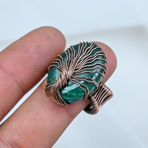Könnte beinhalten: Ein Ring mit einem ovalen, grünen Malachit-Edelstein, der in komplizierten Kupferdraht eingewickelt ist. Der Draht ist in einem verzweigten Muster um den Stein gewickelt und bildet das Band des Rings, was ein metallisches Finish aufweist.