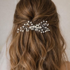 May include: A silver hair comb with pearls and crystals is set in wavy brown hair. The comb is a delicate accessory, perfect for weddings or special occasions. The hair is styled in loose waves.