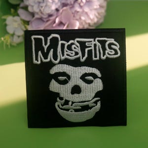 May include: Black square patch with the word "Misfits" embroidered in white above a white skull design. The patch is on a green surface with a colorful lollipop on each side and purple flowers in the background.