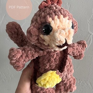 May include: A handmade crochet monkey in shades of mauve and cream, with large black eyes and a yellow banana. The plush toy has a textured, fuzzy appearance. The text "PDF Pattern" is in a pink circle.