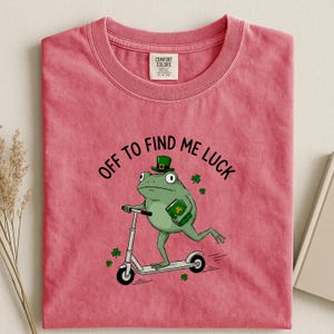 May include: A pink t-shirt with a graphic of a green frog wearing a leprechaun hat and riding a scooter. The frog holds a green book and is surrounded by shamrocks. The text "OFF TO FIND ME LUCK" is above the frog.
