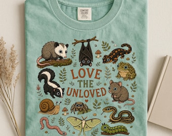 Può includere: Una t-shirt verde salvia con una stampa grafica di vari animali tra cui un opossum, un pipistrello, una salamandra, un rospo, una puzzola, un topo, una lumaca, un ragno, un serpente, una falena, un verme e un bruco con il testo LOVE THE UNLOVED.