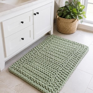 May include: A rectangular, sage green crochet bath mat. The mat is made of thick yarn and has a textured surface. It is placed on a tiled floor in front of a white bathroom vanity.