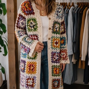 May include: A long, open-front crochet cardigan with a patchwork design of colourful squares. The cardigan has a cream-coloured border and wooden buttons. The garment is worn over a white top and blue jeans. The style is bohemian and handmade.