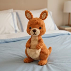 May include: A handmade, crocheted kangaroo plush toy in shades of orange and cream. The kangaroo has large ears, a pouch, and a small tail. It is sitting on a light blue bedspread.