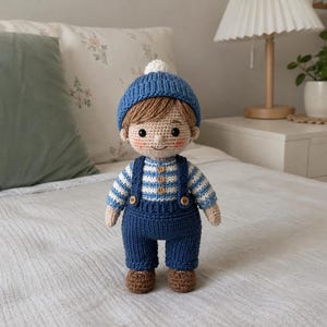 Puede incluir: Muñeca de ganchillo hecha a mano de un niño que lleva un gorro azul tejido con un pompón blanco, una camisa a rayas y un peto azul con zapatos marrones. La muñeca tiene el pelo castaño y una cara sonriente.
