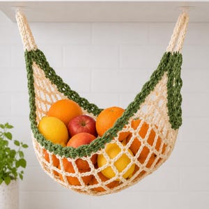 May include: A handmade, cream-colored crochet fruit hammock with a green border, filled with oranges, lemons, and apples. The hammock is suspended from a white surface, offering a stylish and space-saving storage solution for the kitchen.
