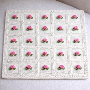 May include: A cream-colored, crocheted blanket featuring a grid pattern with square blocks. Each square is adorned with a pink rose and green leaves. The blanket has a decorative border and is likely handmade.
