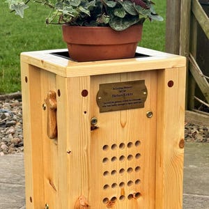 May include: A wooden planter box with a terracotta pot holding a plant with red flowers and green foliage. The box has a decorative wooden handle and a small wooden bird. A brass plaque reads "In Loving Memory MUM Barbara Kirkby".
