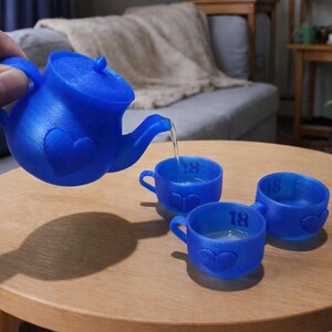 May include: A blue miniature teapot is pouring water into three matching cups. Each cup has a heart design and the number 18. The teapot and cups are on a round wooden table.