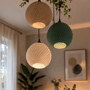 May include: Three pendant lights with a unique, textured design. One is beige, one is green, and one is light beige. The lights are spherical with a spiral pattern and hang from black cords. Greenery hangs from the ceiling above the lights.