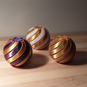 May include: Three decorative spheres with a striped design. One sphere features purple, gold, and dark blue stripes, another has gold and white stripes, and the third is gold. The spheres are on a wooden surface.