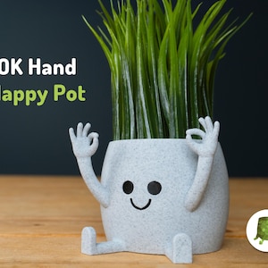 Peut inclure: Un pot de fleurs fantaisiste gris clair en forme de visage souriant, avec des bras et des jambes, faisant le geste de la main "OK". Le pot est rempli d'herbe artificielle verte. Le texte "OK Hand Happy Pot" est visible.