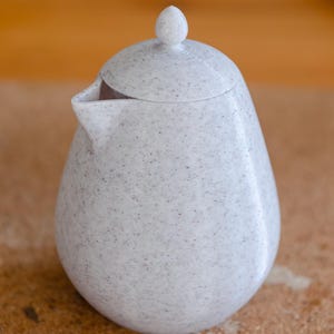 May include: A speckled, off-white ceramic teapot with a lid and a small spout. The teapot has a rounded body and a small, rounded knob on the lid. The surface has a speckled pattern of dark gray and black dots.