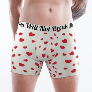 May include: White boxer briefs featuring a repeating pattern of small red hearts. The elastic waistband is white with the black text "You Will Not Break Me" printed in a cursive font across the front centre of the garment.
