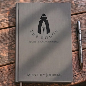 May include: A gray hardcover journal with a black graphic of a hooded figure and a dagger, featuring the text THE ROUGE SECRETS AND CUNNING and MONTHLY JOURNAL. Beside the journal lies a matching gray metal pen with the text SUN & CLERIC.