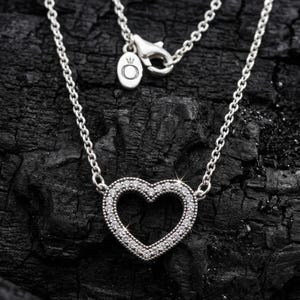 May include: A silver necklace featuring a heart-shaped pendant. The pendant is encrusted with small, sparkling stones. The necklace chain is delicate and has a lobster clasp. Set against a dark, textured backdrop.