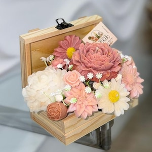 Puede incluir: Una caja de madera llena de un arreglo floral de varias flores en tonos rosa, melocotón y blanco. Las flores están intrincadamente diseñadas, pareciéndose a peonías, margaritas y otras flores. Una pequeña etiqueta está adjunta.