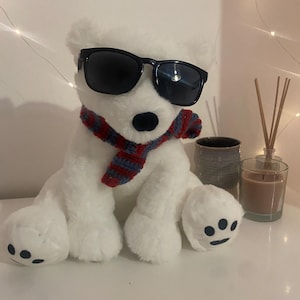 May include: A white plush polar bear wearing black sunglasses and a red, white, and blue striped scarf. The bear has black eyes and paw pads. The bear is sitting on a white surface.