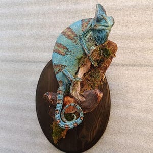 Könnte beinhalten: Eine Wandskulptur eines blauen Chamäleons mit braunen Streifen, das auf einem strukturierten, mit grünem Moos bedeckten Holzstück sitzt. Das gesamte Objekt ist auf einer dunklen, ovalen Holzplatte mit sichtbarer Maserung montiert.