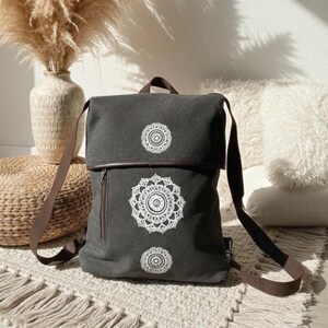 Könnte beinhalten: Ein anthrazitfarbener Rucksack aus Segeltuch mit drei vertikal angeordneten, weißen, kreisförmigen Mandala-Mustern. Der Rucksack verfügt über eine Umschlagklappe, braune Stoffriemen und eine vertikale Reißverschlusstasche auf der linken Vorderseite.