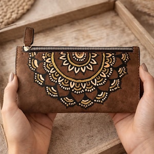 May include: Brown wallet featuring a gold and black mandala design. The wallet has a zip closure and a small pull tab. The design is centrally placed on the front of the wallet.