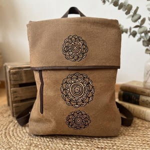 Könnte beinhalten: Ein brauner Rucksack aus Stoff mit Umschlagklappe und drei schwarzen, kreisförmigen Mandala-Mustern, die vertikal in der Mitte aufgedruckt sind. Die Tasche verfügt über eine vertikale Reißverschlusstasche auf der Vorderseite und dunkelbraune Riemen.