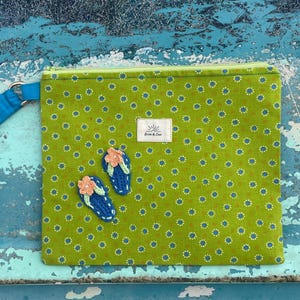 Op de afbeelding: Een groene clutch met patroon en een blauwe polsband. De clutch heeft een bloemenpatroon en twee geborduurde slippers met oranje bloemen. Een klein wit label met tekst staat op de voorkant.