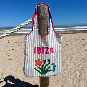 Könnte beinhalten: Eine gestreifte Tragetasche mit dem Wort "IBIZA" in rosa Buchstaben. Die Tasche hat eine rosa Einfassung und Griffe. Auf der Vorderseite sind ein Seestern, Seegras und ein Seepferdchen aufgestickt. Die Tasche hängt an einem Holzpfosten an einem Sandstrand.