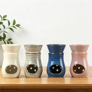 May include: Four ceramic oil warmers in cream, grey, blue, and pink. Each warmer has a bowl on top and a round opening with a dotted design. The warmers are on a wooden surface.