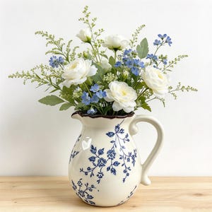 Op de afbeelding: Een keramieken kan met een crèmekleurige basis en een blauw bloemenpatroon, voorzien van een geschulpte rand en een gebogen handvat. De kan is gevuld met een arrangement van witte ranonkels, kleine blauwe bloemetjes en groen blad.