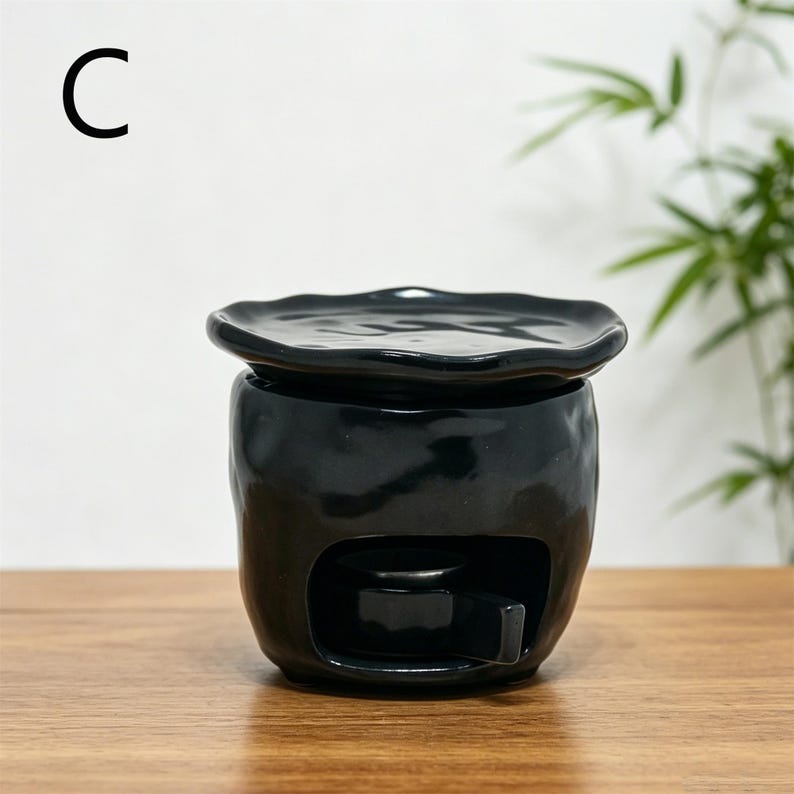May include: A black ceramic essential oil diffuser with a wavy-edged lid. The diffuser has a rounded body with a rectangular opening and sits on a wooden surface. The letter "C" is in the upper left corner.
