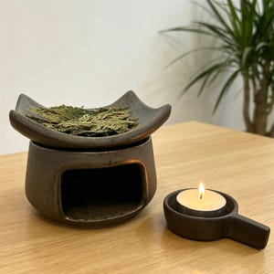 Puede incluir: Un difusor de aceites esenciales de cerámica marrón oscuro con una parte superior cuadrada que contiene hojas secas. Un pequeño portavelas redondo con una vela encendida está al lado del difusor. Los objetos están sobre una superficie de madera.