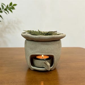 May include: A ceramic essential oil diffuser with a lit tea light candle inside. The diffuser is beige with a small dish on top holding green dried leaves. The candle flame glows brightly, creating a warm ambiance.