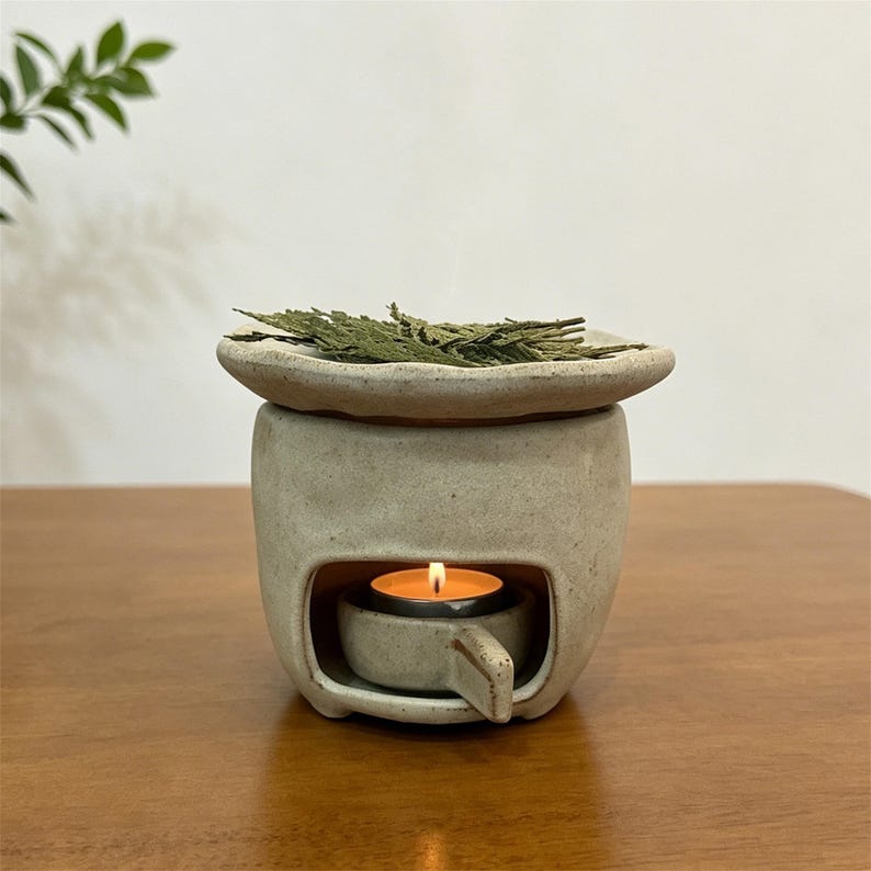 May include: A ceramic essential oil diffuser with a lit tea light candle inside. The diffuser is beige with a small dish on top holding green dried leaves. The candle flame glows brightly, creating a warm ambiance.