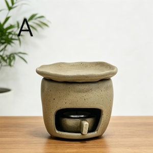 May include: A beige ceramic essential oil diffuser with a small, round opening for a tea light. The diffuser has a matching lid and is sitting on a wooden surface. The letter "A" is in the upper left corner.