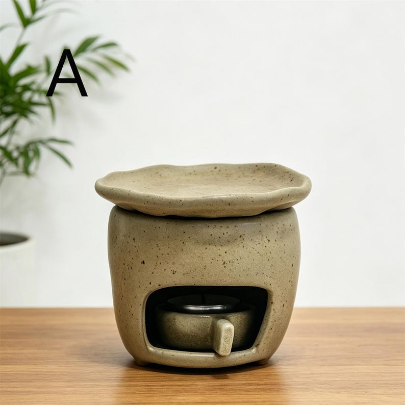 May include: A beige ceramic essential oil diffuser with a small, round opening for a tea light. The diffuser has a matching lid and is sitting on a wooden surface. The letter "A" is in the upper left corner.