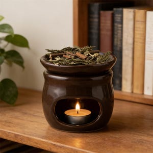 May include: A brown ceramic oil warmer with a lit tealight candle inside. The top dish holds dried herbs and spices. The warmer sits on a wooden surface, with a bookshelf in the background.