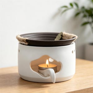 May include: A white ceramic essential oil diffuser with a dark brown dish and a lit tea light candle. The diffuser has a leaf-shaped cutout and a jute rope detail. The candle flame is visible.