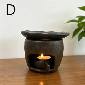 May include: A black ceramic essential oil diffuser with a lit tea light candle inside. The diffuser has a small, wavy-edged dish on top and a rectangular opening in the front. The letter "D" is in the upper left corner.