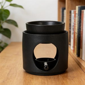 May include: A black ceramic essential oil warmer with a textured finish. It features a circular opening and a small dish for oil. A small dish is inside the opening to hold a tea light. The warmer is on a wooden surface.