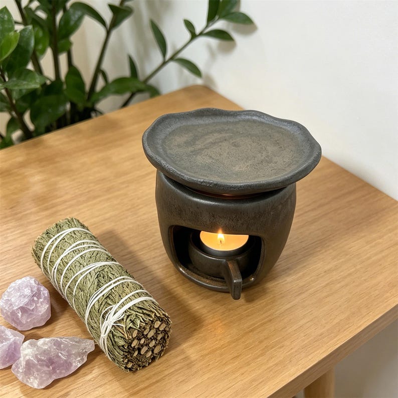 May include: A dark gray ceramic essential oil diffuser with a lit candle inside, a sage bundle, and amethyst crystals on a wooden surface. The diffuser has a round base and a wavy-edged top. The sage bundle is wrapped with white string.
