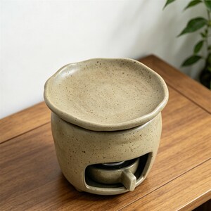 May include: A beige ceramic essential oil warmer with a speckled finish. The warmer has a round base with a small opening and a removable dish on top. The design is simple and modern, suitable for aromatherapy and home fragrance.